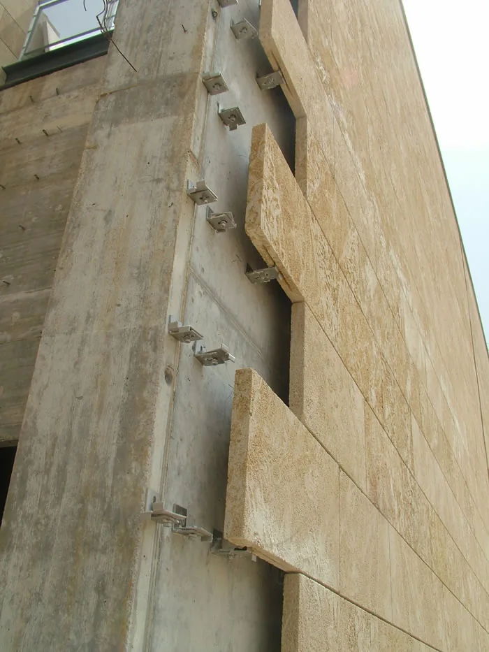 Ventilated natural-stone facade under installation: stainless edge clips bolted to the backup wall, engaging the kerf on the top and bottom edge of each stone panel inside the open joint, with nothing fixed through the face