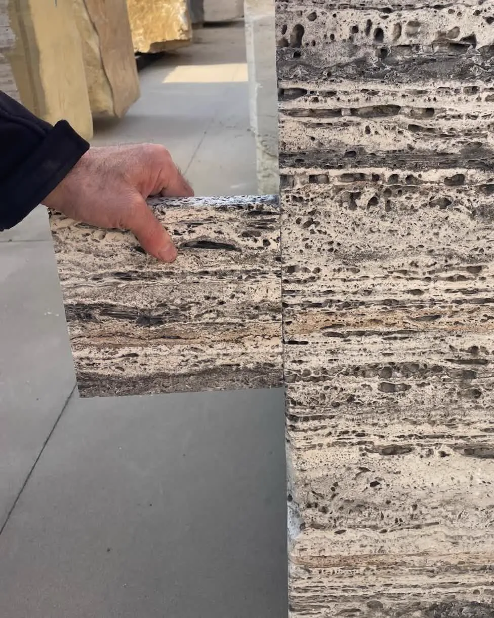 Honed travertine sample held against the sawn face of the block to check color and texture, block yard behind