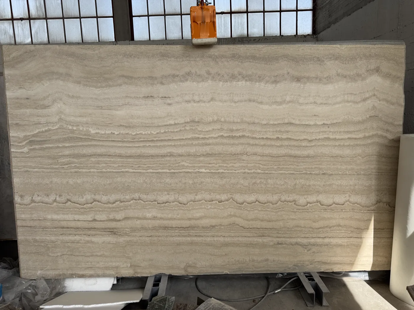 Vein-cut travertine slab hanging from an overhead clamp lifter in the workshop