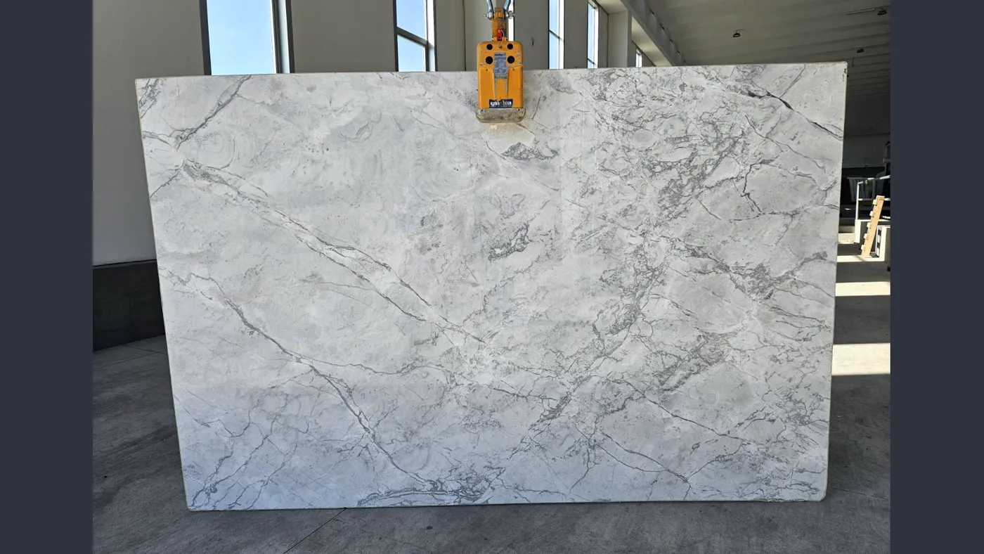Full white marble slab standing in the warehouse during selection, lifting clamp resting on the top edge
